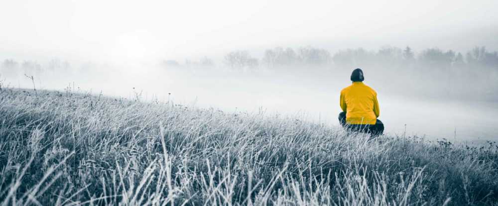 Foto av en åker i frost. Til høyre for bildet, omtrent i midten dybdemessig, sitter enmann på huk og ser videre inn i bildet. Han ser på en tåkebelagt åker og en skoglinje i det fjerne. Fotoet er i blågrå toner. Unntaket er jakken til mannen, som er i en sterk guloransje farge.