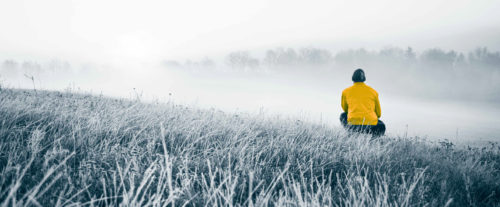 Foto av en åker i frost. Til høyre for bildet, omtrent i midten dybdemessig, sitter enmann på huk og ser videre inn i bildet. Han ser på en tåkebelagt åker og en skoglinje i det fjerne. Fotoet er i blågrå toner. Unntaket er jakken til mannen, som er i en sterk guloransje farge.
