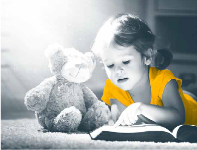 Halvnært foto av en liten jente som ligger på gulvet og peker i en oppslått bok som ligger på gulvet. Til høyre fpr henne er det en teddybjørn som sitter på gulvet. Bakgrunnen er uklar og hele fotoet er i gråtoner. Unntaket er jentas bluse, som er i en guloransje farge.