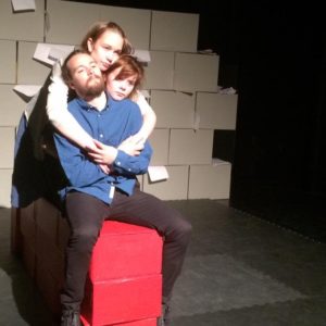 Foto av en teaterscene. På en rød mur laget av røde klosser (tre i høyden og fem i bredden) sitter tre mennesker: En mann med olaskjorte, sort bukse og skjegg og en kvinne med brunt hår og panenlugg hvis hode titter frem over mannens skulder. Bak kvinnen sitter en andre kvinne og omfavner begge de to foran seg. Gulvet er sort. I bakgrunnen ser vi en vegg satt opp av grå pappesker.