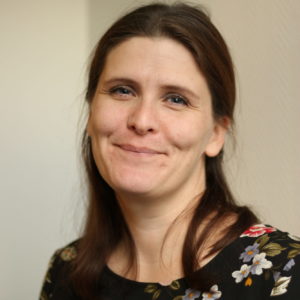 Foto av kvinne med mørkebrunt hår bak ørene og til skuldrene. Hun smiler og ser inn i kameraet med hodet litt på skakke. Hun har en sort bluse med blomsterprint på. Bakgrunnen er en hvit vegg.