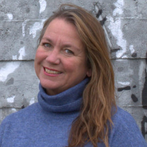 Foto av kvinne som smiler bredt og ser inn i kamera. Hun har lysebrunt/mellomblondt hår til skuldrene, blå øyne og en pologenser i en tåkeblå farge. Bakgrunnen er en røff betongvegg i grå med hvite og sorte malingsflekker på. Fotoet er tatt fra litt under skuldrene og opp.