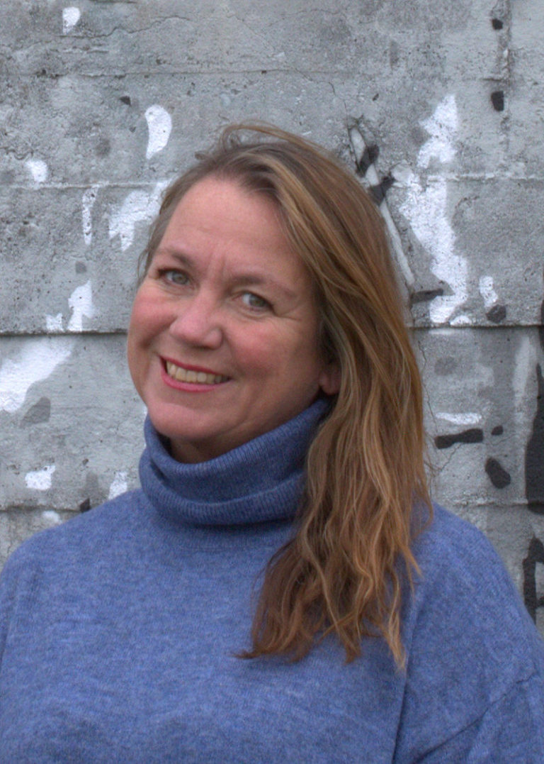 Gro Harestad 2 Foto av kvinne som smiler bredt og ser inn i kamera. Hun har lysebrunt/mellomblondt hår til skuldrene, blå øyne og en pologenser i en tåkeblå farge. Bakgrunnen er en røff betongvegg i grå med hvite og sorte malingsflekker på. Fotoet er tatt fra litt under skuldrene og opp.