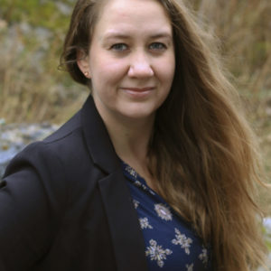 Kvinne med langt hår til bysten ser rett inn i kameraet og smiler. Hun har på seg en sort dressjake over en mørkeblå bluse med mønstertrykk. Kvinnen har blå øyne og lys gud. Bakgrunnen er uklar natur.