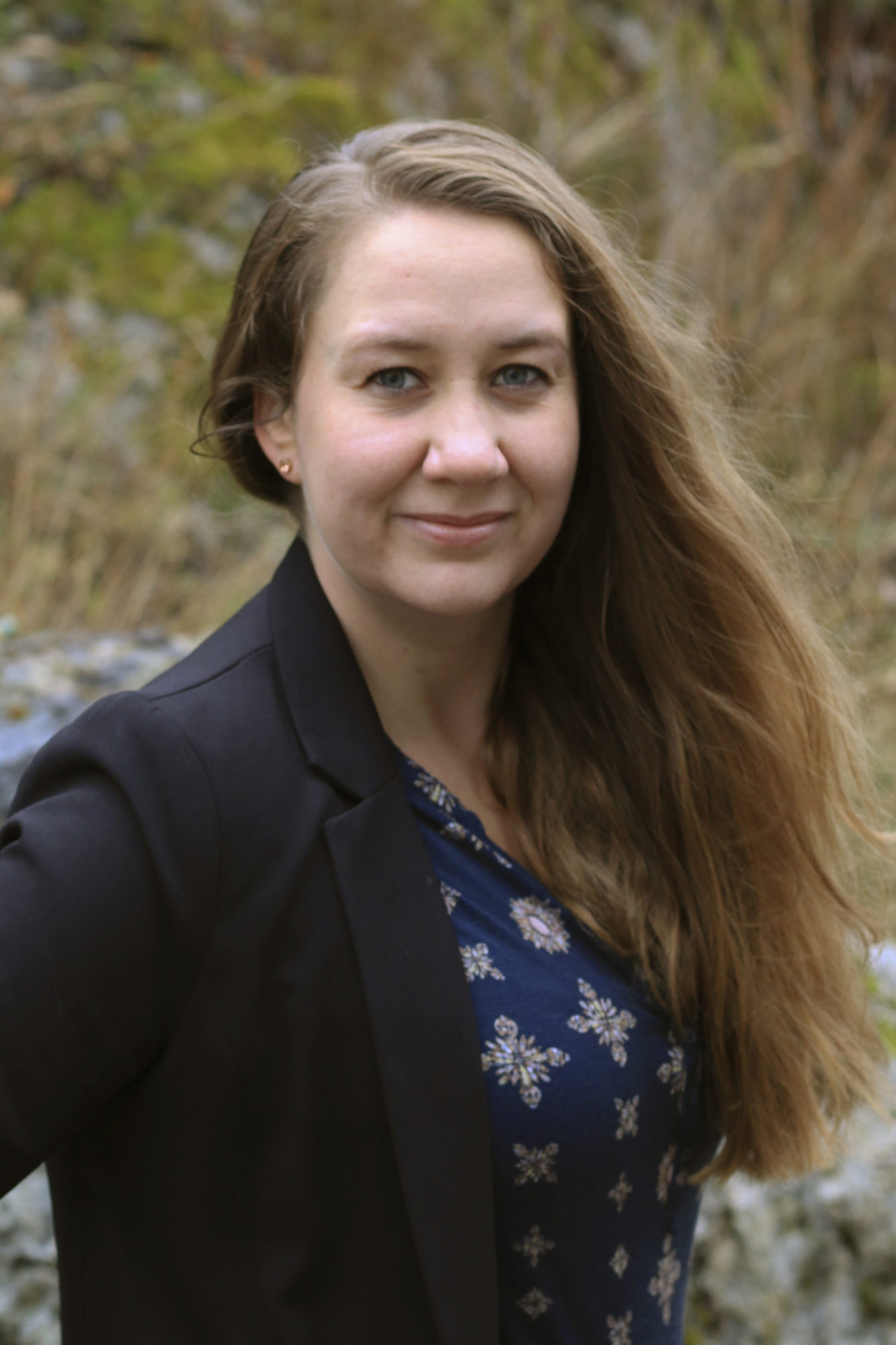 Kvinne med langt hår til bysten ser rett inn i kameraet og smiler. Hun har på seg en sort dressjake over en mørkeblå bluse med mønstertrykk. Kvinnen har blå øyne og lys gud. Bakgrunnen er uklar natur.