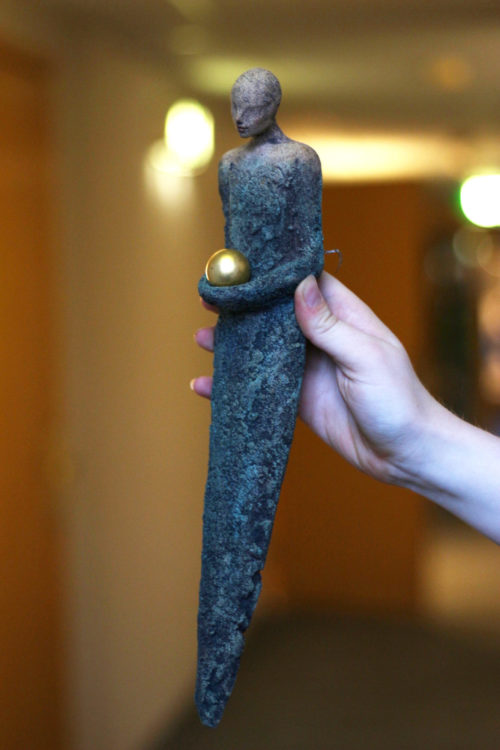 Fotografi av en skulptur. Skulpturen er en menneskelignende figur med menneskelignende torso og to ben som er smeltet sammen i et, så det ligner et skjørt som ender i en traktform. Personen holder armene smeltet sammen rundt en kule av gull på midten av kroppen.