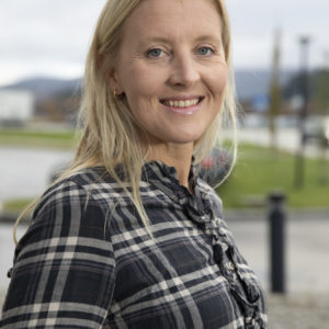 Foto av kvinne med blondt hår til litt lenger enn skuldrene. Hun har på seg en rutete skjorte og smiler og ser nn i kameraet. Hun har blå øyne. Bakgrunnen er et uklart landskap.