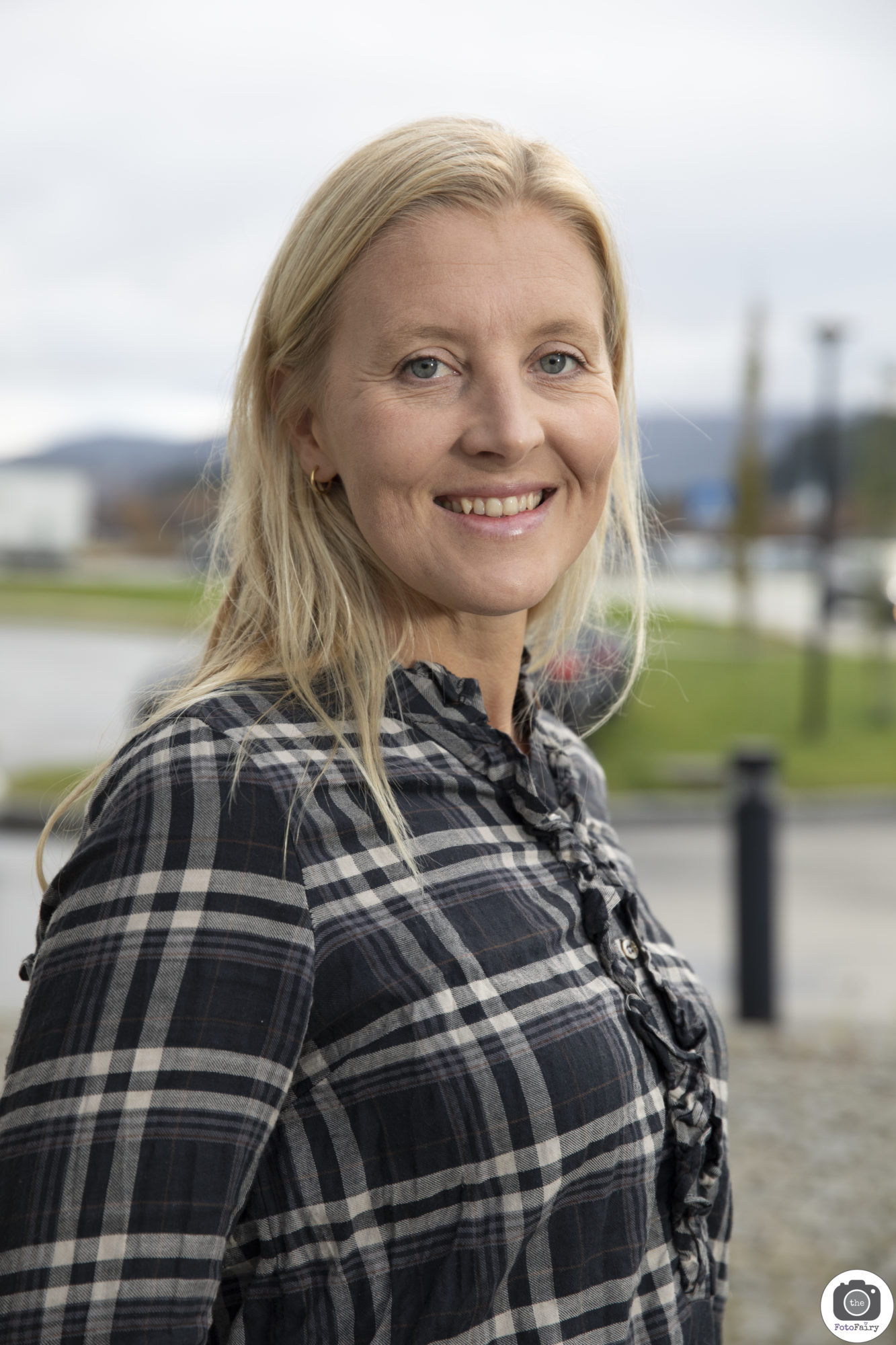 Foto av kvinne med blondt hår til litt lenger enn skuldrene. Hun har på seg en rutete skjorte og smiler og ser nn i kameraet. Hun har blå øyne. Bakgrunnen er et uklart landskap.