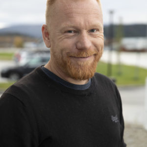 Foto fra livet og opp av mann med skjegg, blå øyne og rødt hår, Han ser inn i kameraet og smiler. Bakgrunne er et uklart landskap.