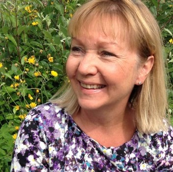 Voksen kvinne med blondt hår til skuldren, pannelugg og hvit bluse med lilla blomstermønster ser tl høyre i bildet og smiler. Bildet er tatt i halvprofil. I bakgrunnen er det grønt gress med gule blomster. Bildet er tatt fra overarmene og opp.
