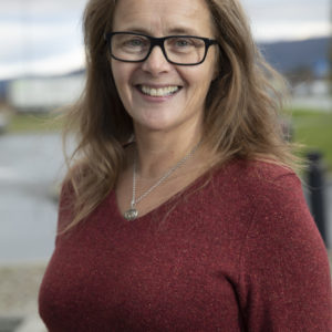 Foto av kvinne med mørkeblondt hår til litt under skuldrene. Hun har briller og smiler bredt mens hun ser rett inn i kamera. Hun har på seg en burgunder genser. Bakgrunnen er et uklart landskap.