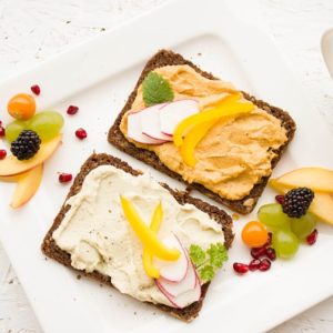 Foto av to knekkebrød på en hvit, firkantet asjett. På knekkebrødene er det påsmurt hummus. På asjetten er forskjellig frukt som bjørnebær, druer og skivede epler. Bildet er tatt rett ovenfra.