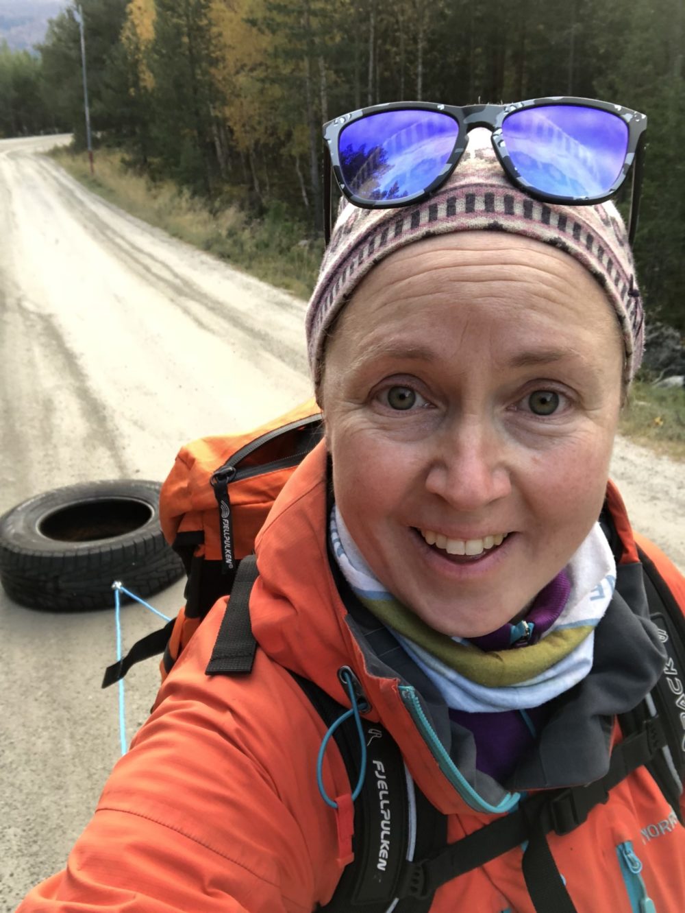 En kvinne med brune øyne, oransje treningsjakke, lue og solbriller i panna/på hodet ser inn i kameraet og smiler. Bak henne ser vi at hun trekker på et bilhjul på en skogsvei.