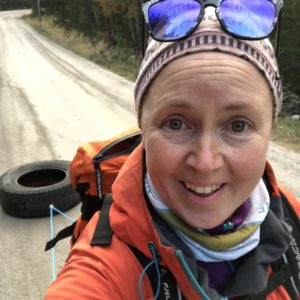En kvinne med brune øyne, oransje treningsjakke, lue og solbriller i panna/på hodet ser inn i kameraet og smiler. Bak henne ser vi at hun trekker på et bilhjul på en skogsvei.