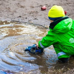 En liten gutt sitter med ryggen til i grønn termodress. Han leker med en bil som han kjører i en sølepytt.
