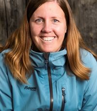 En kvinne med gyllenbrunt hår til litt lenger enn skuldrene ser inn i kameraet og smiler bredt. Hun har på seg en lyseblå utendørsjakke. Bakgrunnen er uklar natur. Fotoet er fra overarmene og opp.