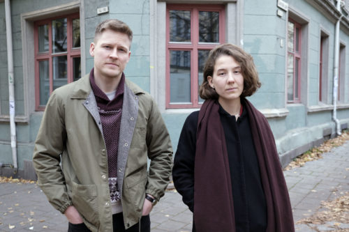 En mann og en kvinne står foran et bygg og ser inn i kameraet. Mannen har grågrønn jakke og kortklipt hår. Kvinnen har sort jakke og lilla skjerf. De har en nøyral mine og ser rett i kamera. Bakgrunnen er et grønt bygg med vinduer med røde sprosser.