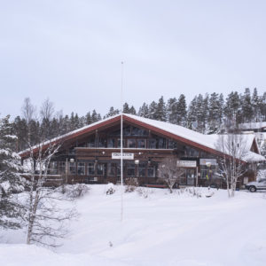 Foto av en stor fjellhytte med skrånende tak omgitt av trær og snø.
