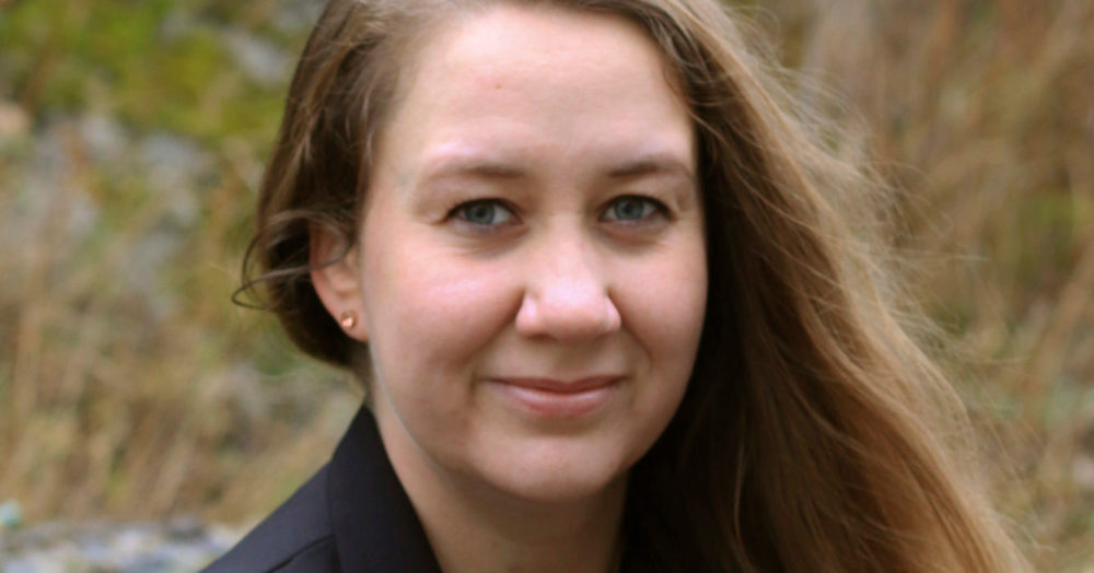 En kvinne smiler og ser inn i kameraet. Hun har langt, brunt hår. I bakgrunnen er det uklar natur.