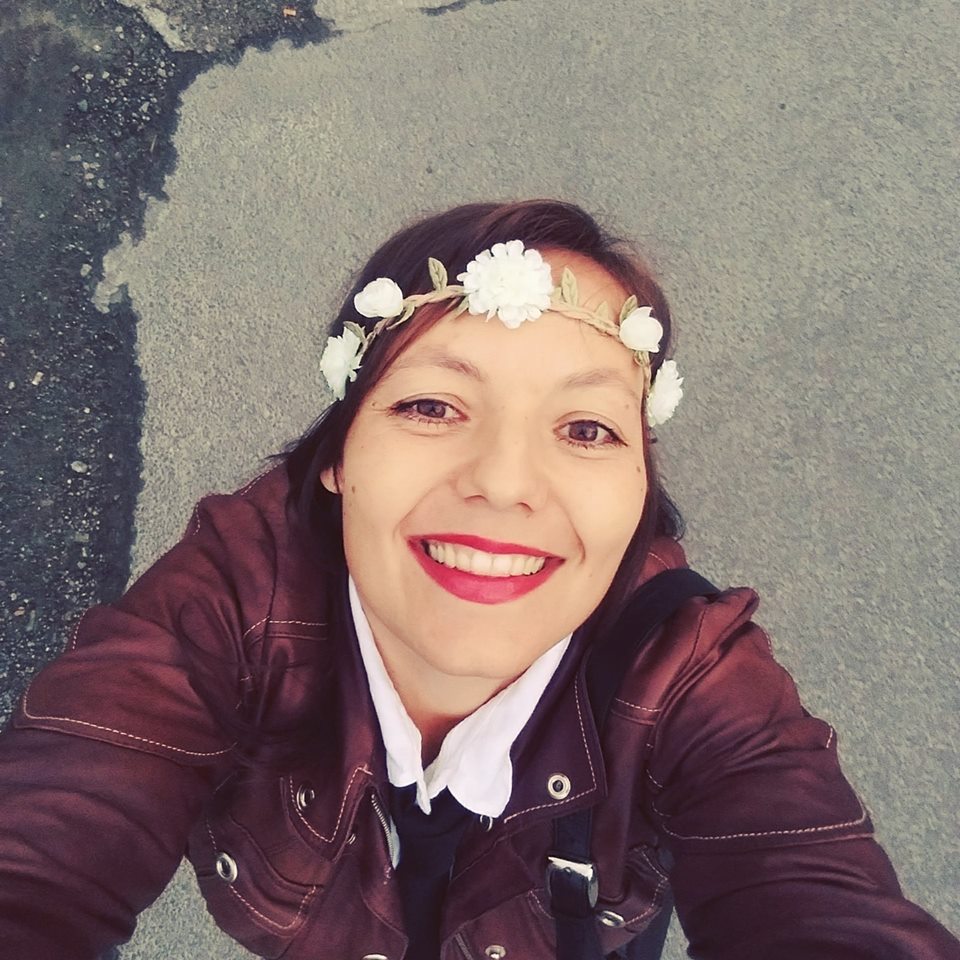 Selfie av kvinne ovenfra. Kvinenn har langt, mørkt hår, brugunder jakke og hvit skjorte med sort genser over. Hun har på seg et pannebånd/strikk med hvite blomster i krans rundt hodet. Hun smiler og ser inn i kamera. Bak henne ser vi asfalt.