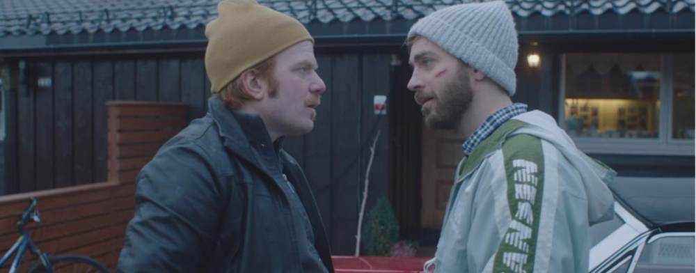 Foto av to menn som står ansikt til ansikt i profil. Bak dem er en husvegg. Begge har på seg ytterklær og lue. De ser opprørte ut. Mannen til høyre har grå ytterklær og brunt skjegg og grå lue. Mannen til venstre har oranasje hår, gul lue og grønn jakke.