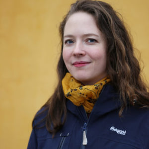Kvinne med lys hud og mørkebrunt hår ser inn i kameraet og smiler med munnen lukket. Hun har på seg et gult skjerf og blå Bergans-jakke. I bakgrunnen er en okergul vegg.