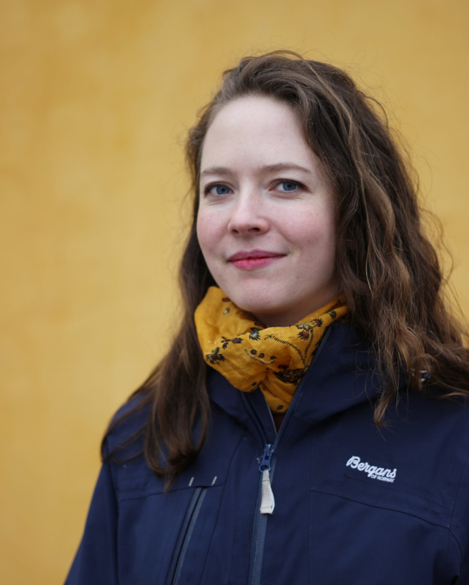 Kvinne med lys hud og mørkebrunt hår ser inn i kameraet og smiler med munnen lukket. Hun har på seg et gult skjerf og blå Bergans-jakke. I bakgrunnen er en okergul vegg.