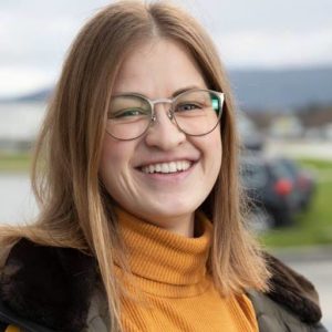 Foto av kvinne som ser rett i kamera. Hun har mørkblondt hår til skuldrene, oransje genser, brun ytterjakke og briller. Hun smiler. Bakgrnnen er uklar med natur og en parkeringsplass.