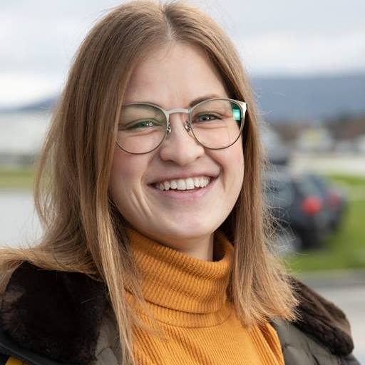Foto av kvinne som ser rett i kamera. Hun har mørkblondt hår til skuldrene, oransje genser, brun ytterjakke og briller. Hun smiler. Bakgrnnen er uklar med natur og en parkeringsplass.
