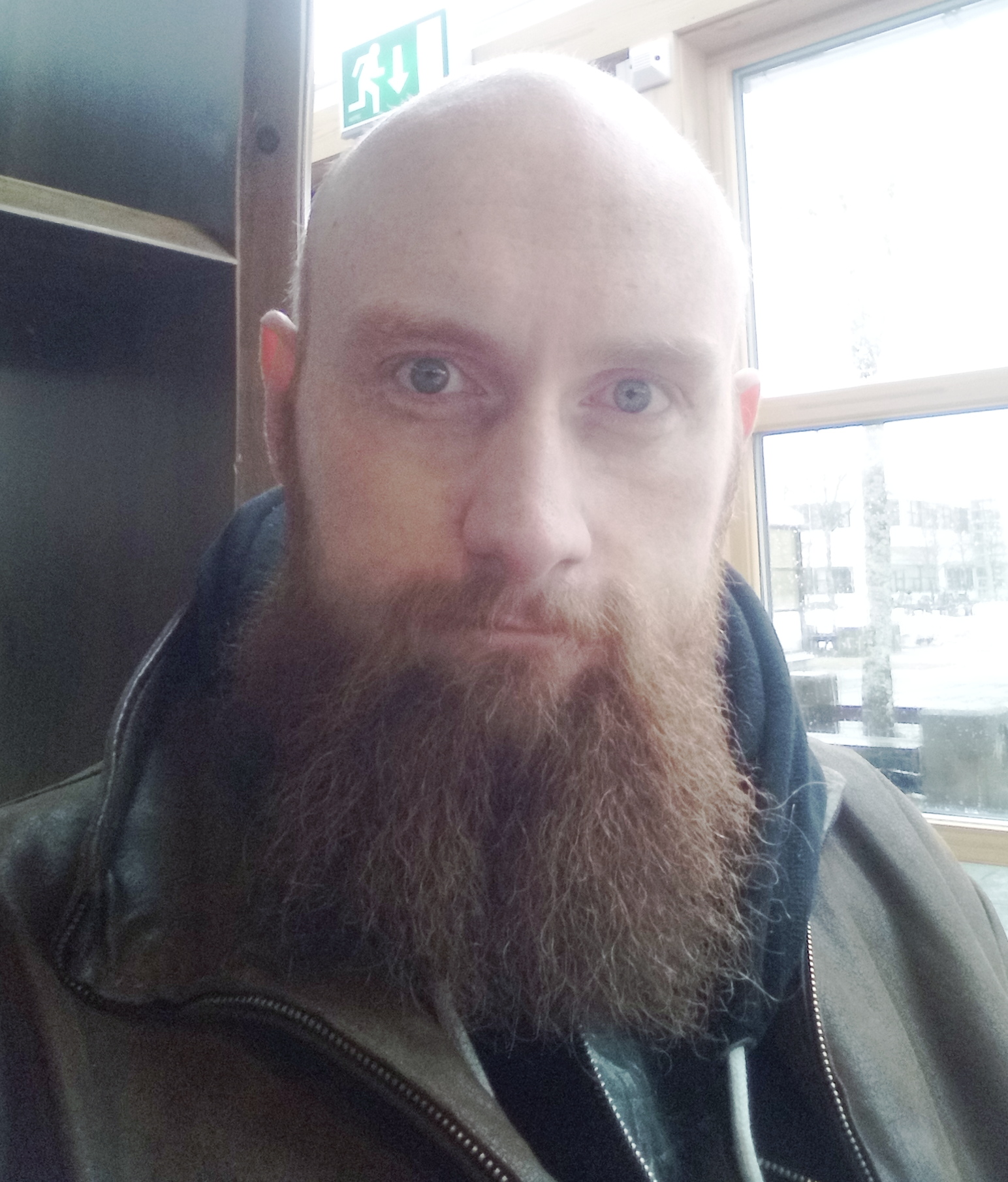 Foto av mann som ser inn i kameraet. Han har glattbarbert hode, kraftig rødlig skjegg og blå øyne. Han har på seg en hettegenser og en skinnjakke.