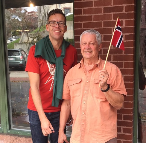 Foto av to menn som ser i kameraet og smiler. Vi ser den fra livet og opp. Mannen til venstre har rød t-skjorte, en genser knytt om halsen, briller og kort, brunt hår. Mannen til høyre har en lys lakserosa skjorte, grått, kort hår og holder et norsk falgg i hånden. I bakgrunnen er en murvegg med rød murstein.