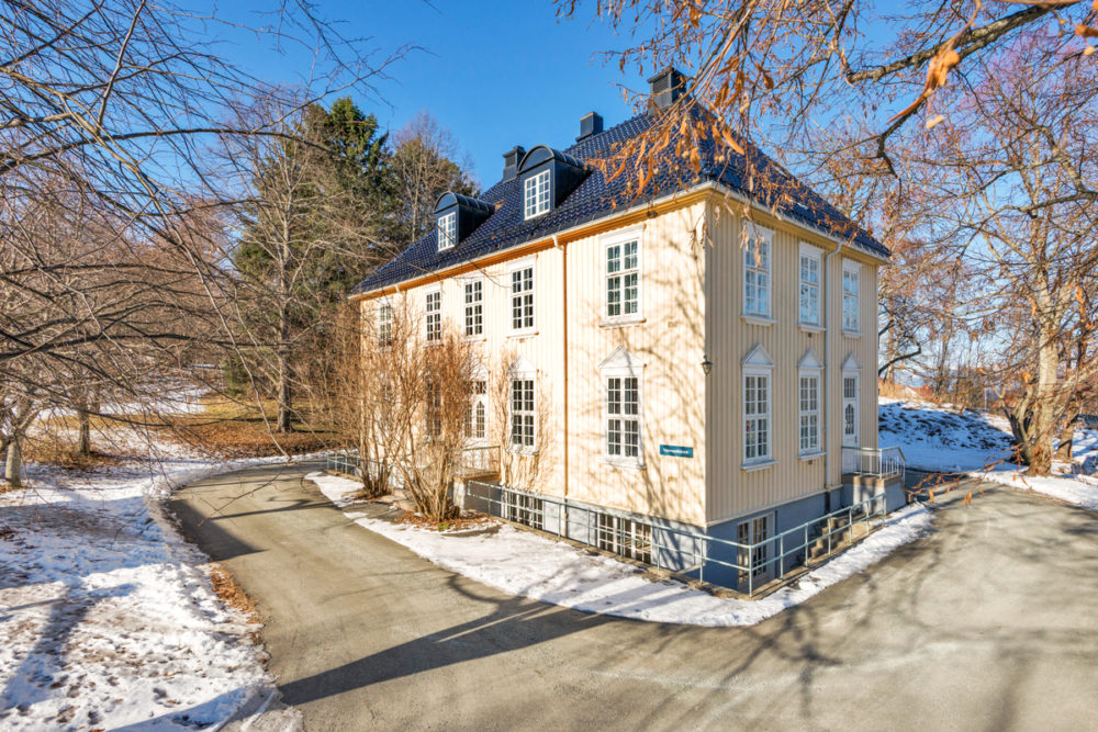 Foto av et bygg malt i lys gul. Det er sprosser på vinduene, og ellers gammeldags stil. Det er blå himmel og sol og litt snø på omgivelsene.