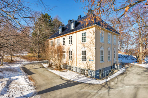 Foto av et bygg malt i lys gul. Det er sprosser på vinduene, og ellers gammeldags stil. Det er blå himmel og sol og litt snø på omgivelsene.