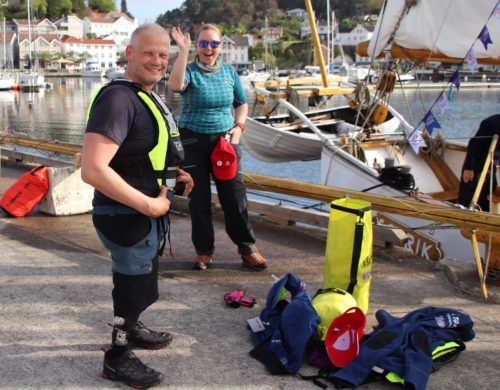 Foto av en kvinne og en mann ytterst på en havn. Kvinen har blå skjorte, solbriller og vinker til kamera. Mannen har sort t-skjorte, livvest og proteser på beina. Foran dem står en haug med diverse utstyr de skal bruke i sammenheng med toktet: Sovepose, ting i pressening osv.