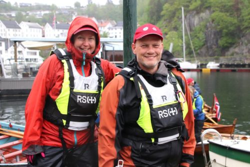 Foto av kvinne til venstre og mann til høyre i oransje rengjakke og gul og sort flytevest. DE har rød caps. Bak dem er en havn med båter. Det regner.