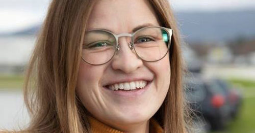 Foto av kvinne som ser rett i kamera. Hun har mørkblondt hår til skuldrene, oransje genser, brun ytterjakke og briller. Hun smiler. Bakgrnnen er uklar med natur og en parkeringsplass.