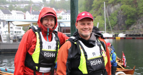 Foto av kvinne til venstre og mann til høyre i oransje rengjakke og gul og sort flytevest. DE har rød caps. Bak dem er en havn med båter. Det regner.