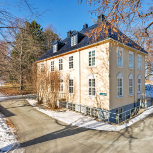 Foto av et bygg malt i lys gul. Det er sprosser på vinduene, og ellers gammeldags stil. Det er blå himmel og sol og litt snø på omgivelsene.