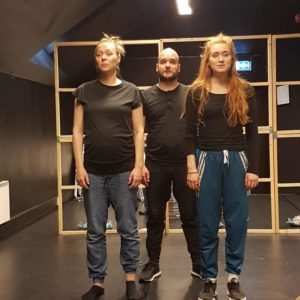 Foto av tre personer med mørke bukser og t-skjorter/gensere som står rett opp og ned, tett i tett. Det er en kvinne, en mann i midten, og så en kvinne. De står i et rom med sorte vegger og foran en skillevegg av tre.